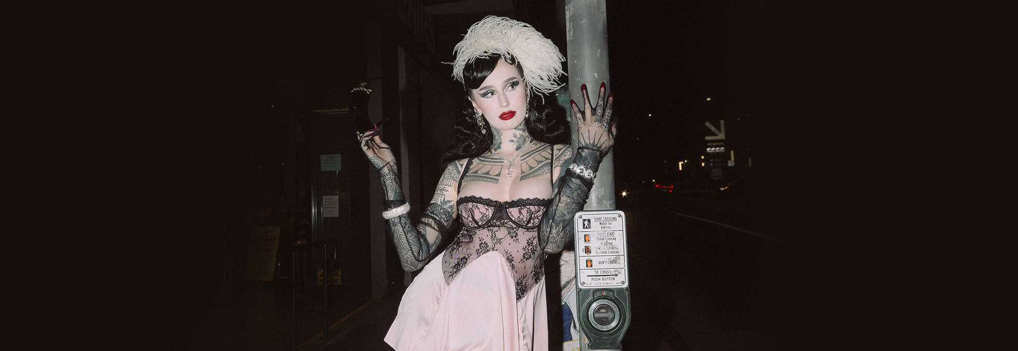 A person with tattoos, dramatic makeup, and a feathered headpiece poses at night in a sheer lace dress with gloves, holding a cigarette next to a parking meter on a city street.