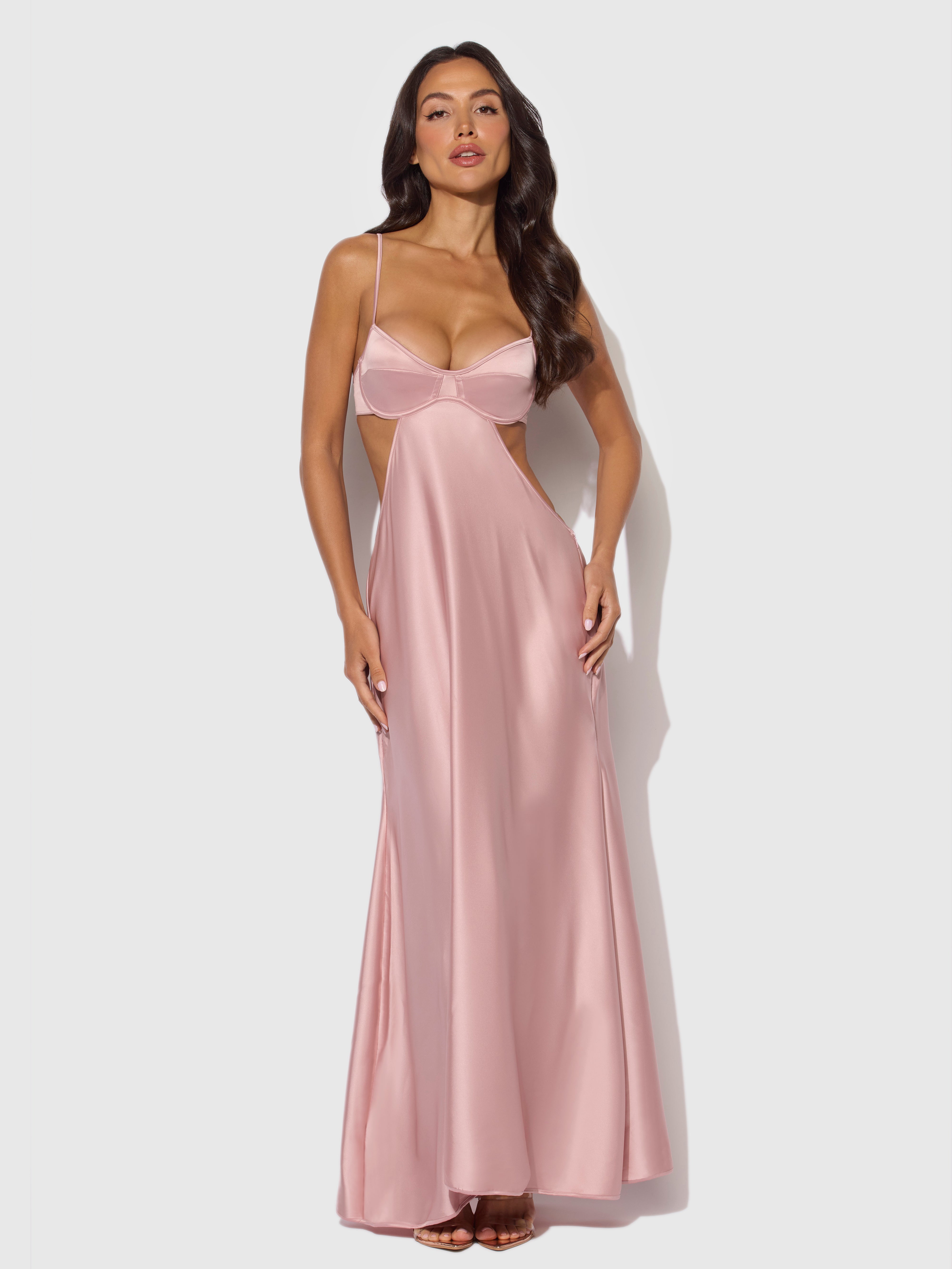 A woman stands against a plain white background wearing the Frederick's of Hollywood Petra Satin Monowire Cutout Gown, a long, silky light pink dress featuring thin straps, cutouts, and supportive satin monowire detailing.