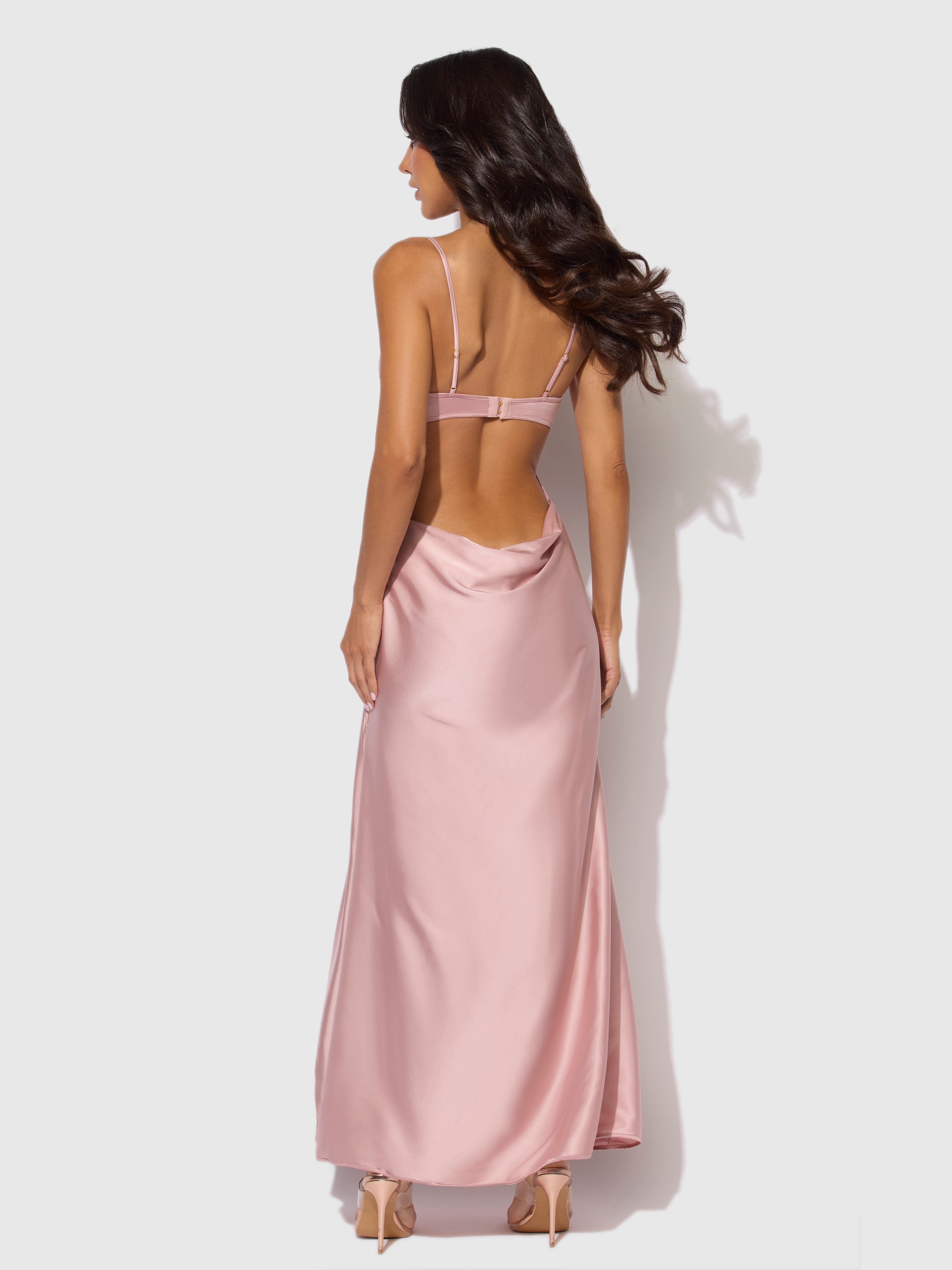 A woman with long, wavy dark hair models the Frederick's of Hollywood Petra Satin Monowire Cutout Gown—backless, silky pink with thin straps—paired with nude heels, posing against a plain light background.