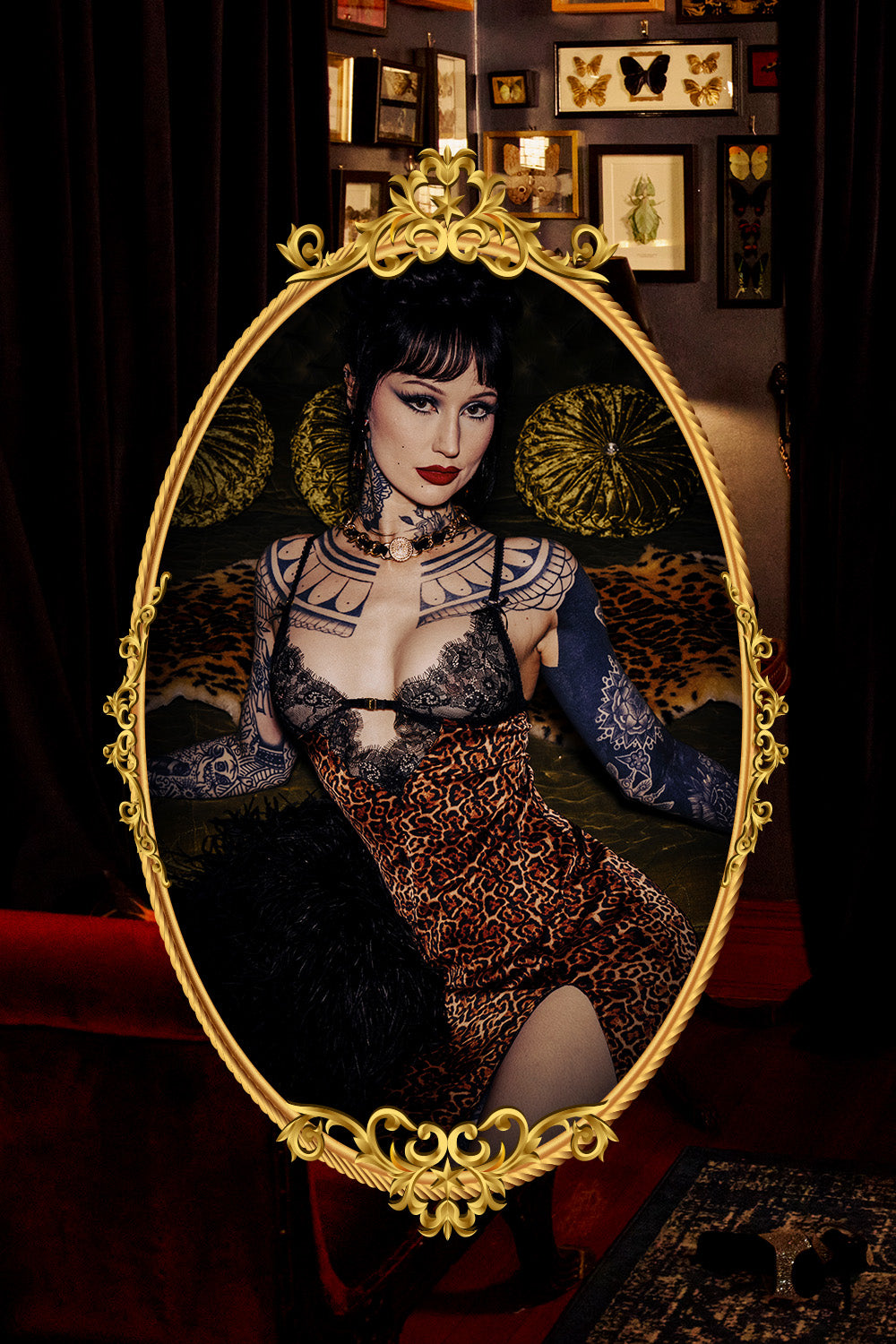 A gothic-style portrait in an ornate gold oval mirror frame, showing a woman with dark hair, red lipstick, and tattoos wearing a leopard-print dress and lace gloves, posed in a dark room with framed wall art and red velvet seating.