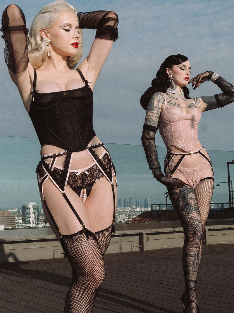 Two women pose confidently on a rooftop, one wearing Frederick's of Hollywood's Rosalind Lace & Pearl Crotchless Thong in black with a removable pearl strand, the other in pink. Stockings and gloves complete their vintage-inspired looks against the city skyline.