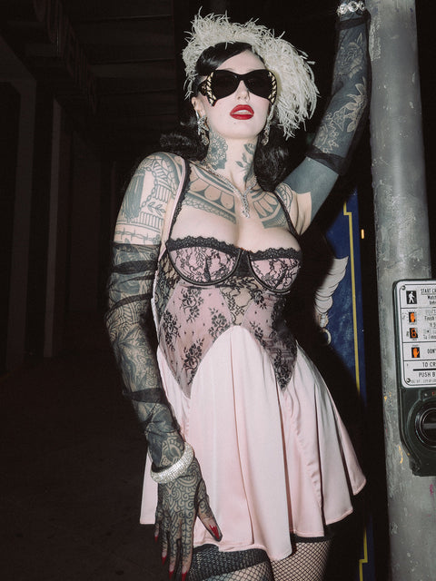 A tattooed person in black sunglasses, a feathered headpiece, black lace gloves, and the Frederick's of Hollywood Rosalind Lace Bodice & Satin Skirt Babydoll poses outdoors at night with one arm resting on a pole.