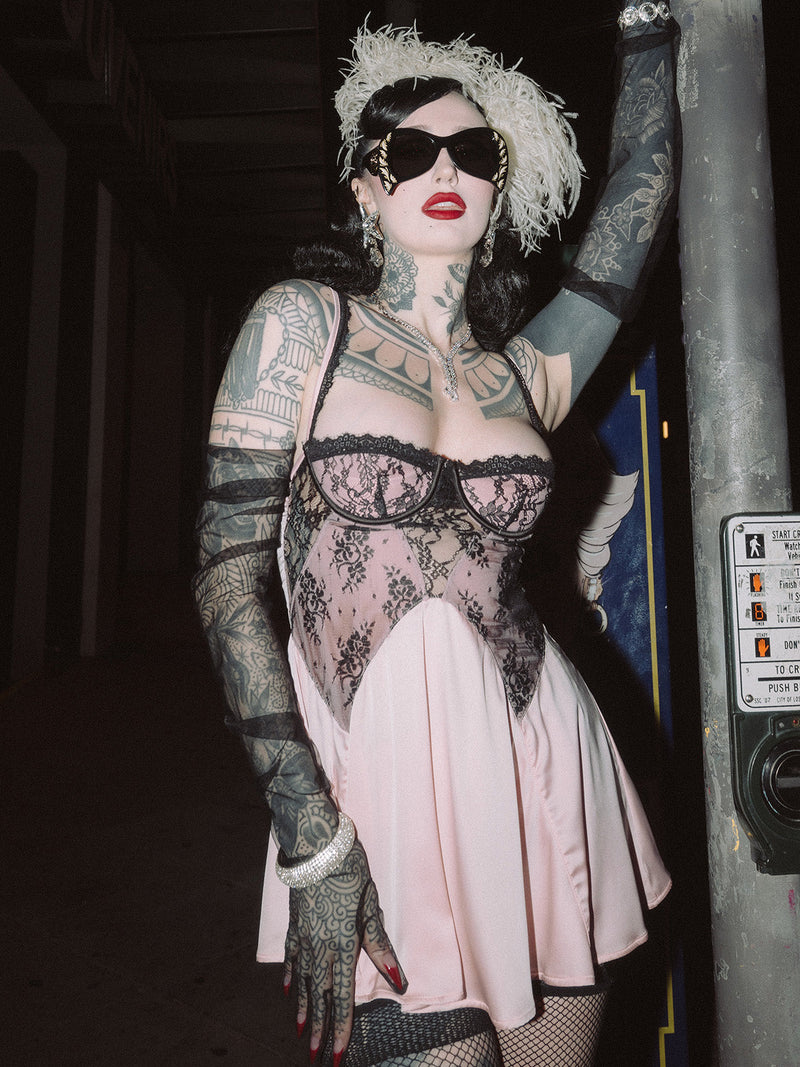 A tattooed person in black sunglasses, a feathered headpiece, black lace gloves, and the Frederick's of Hollywood Rosalind Lace Bodice & Satin Skirt Babydoll poses outdoors at night with one arm resting on a pole.