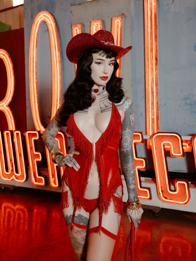 A woman with long dark hair and tattooed arms poses confidently in the Hendrix Faux Leather & Fringe Chemise by Frederick's of Hollywood, paired with a red cowgirl hat and gloves, standing before large neon signs.
