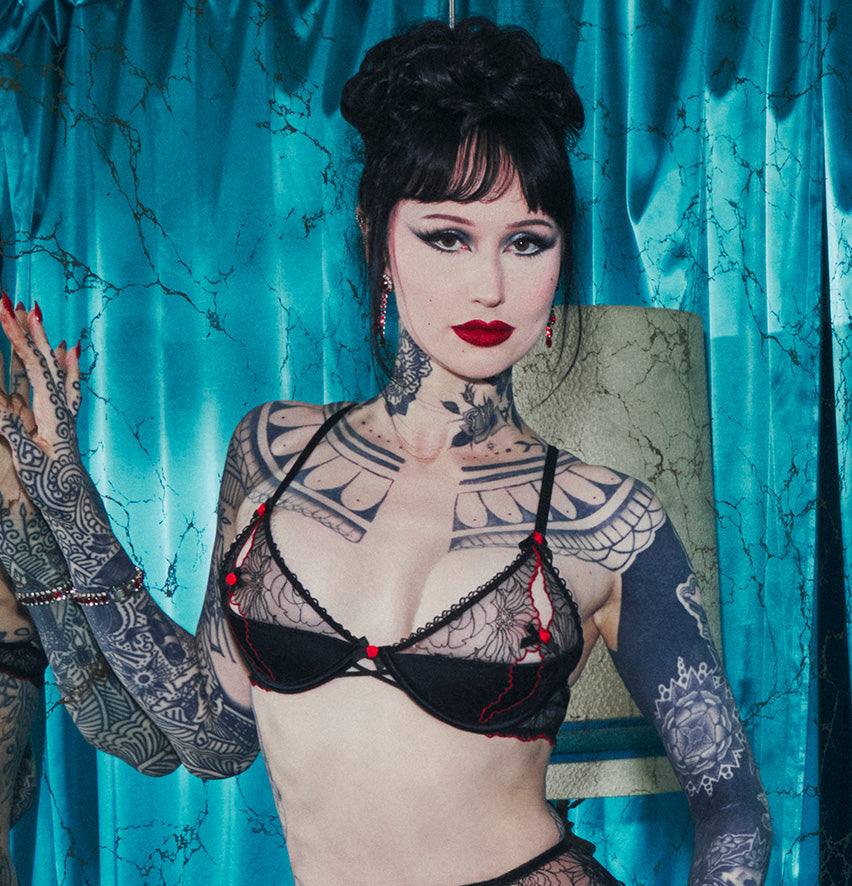 A tattooed woman with dark hair styled in a high bun poses in front of a textured turquoise curtain, wearing a black lace bralette with red accents.