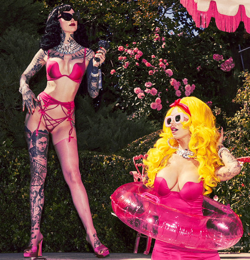 Two women in pink outfits with tattoos and yellow hair in a garden setting.