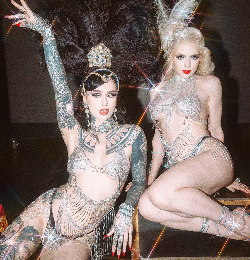 Two women in glamorous showgirl costumes with sparkling silver fringe, elaborate feathered headdresses, and bold makeup pose together under bright, glittering lights, evoking a vintage cabaret style.