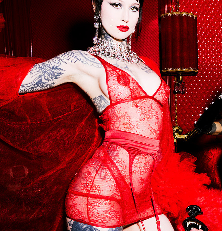 A woman modeling a red lace lingerie bodysuit with garter straps and a matching red sash, posed against a dark red velvet backdrop with a red lamp and decorative setting visible.