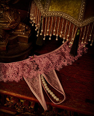 A pink lace garter belt with matching ribbons and pearl accents, displayed on a dark wooden surface next to a decorative gold and brown fringed textile.