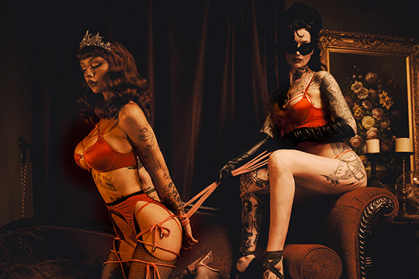 Two tattooed women pose in revealing orange lingerie and strappy harnesses in a dark, moody room with a sofa, curtains, and a framed floral painting.