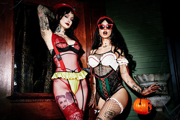 Two women posing in Halloween costumes in a dimly lit house setting. The woman on the left wears a red and black lingerie-style costume with a yellow ruffle and thigh-high stockings.