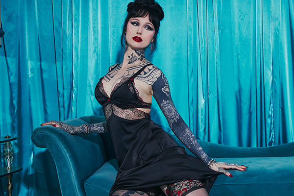 A woman with dark hair and visible tattoos poses on a teal velvet sofa in front of a matching teal curtain backdrop, wearing a black lace and satin dress.