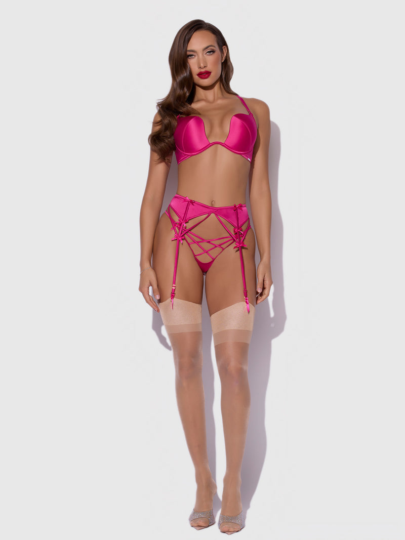 A woman poses confidently against a plain background wearing Frederick's of Hollywood's Petra Satin & Mesh Garter Belt in hot pink, styled with a matching bra and beige stockings. She has long brown hair and red lipstick.