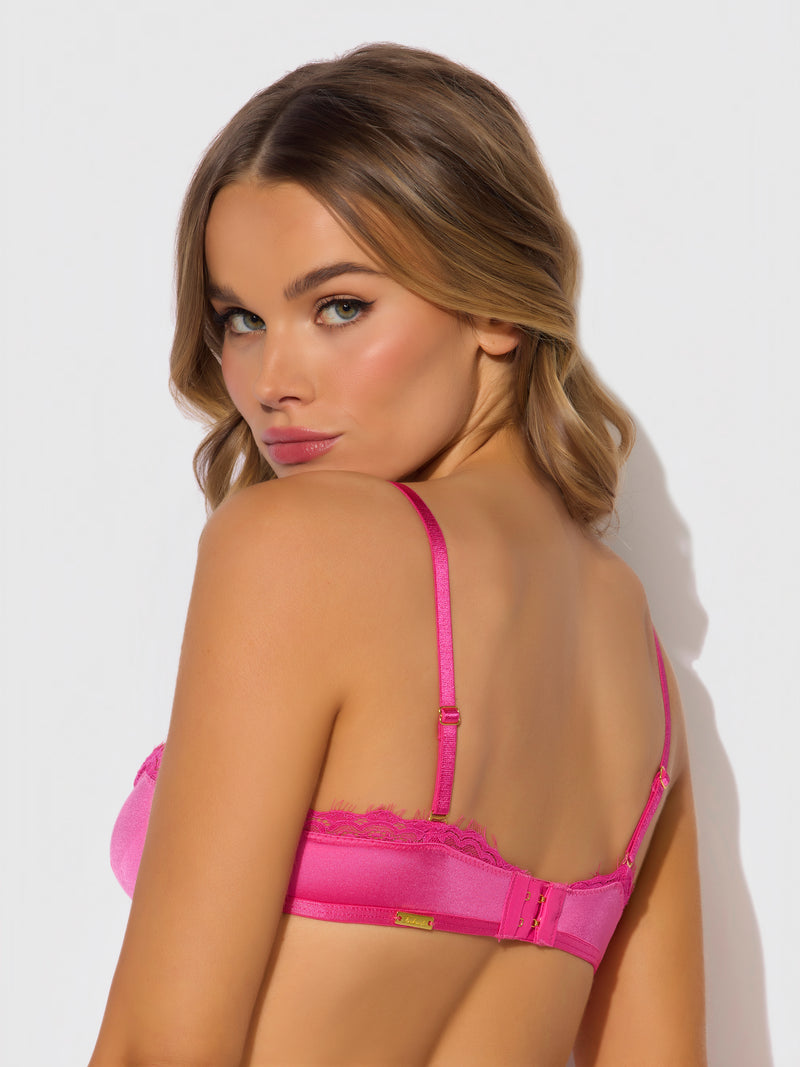 A woman with wavy, light brown hair looks over her shoulder while wearing the Frederick's of Hollywood Neev Lace & Oilslick Push Up Half Up Bandeau Bra in bright pink, posing against a plain, light background.