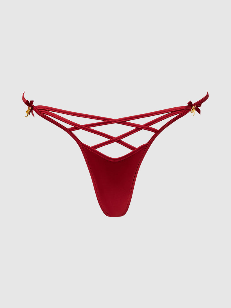 The Frederick's of Hollywood Petra Heart Open Back Bikini is a red, low-cut strappy thong with multiple crisscrossing front straps and gold bow accents on each hip, shown on a plain light gray background.