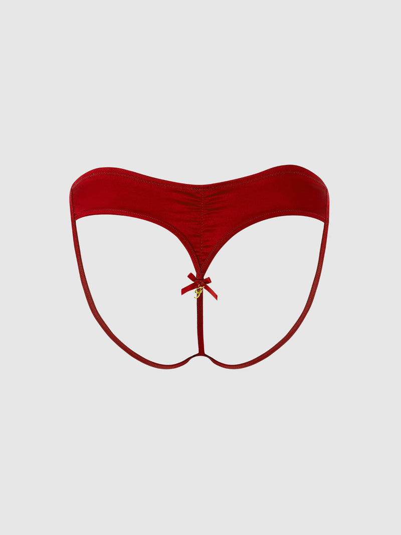 The Petra Heart Open Back Bikini by Frederick's of Hollywood is a red thong featuring a thin waistband, ruched center seam, and a small gold charm and bow at the front, shown against a plain light gray background.