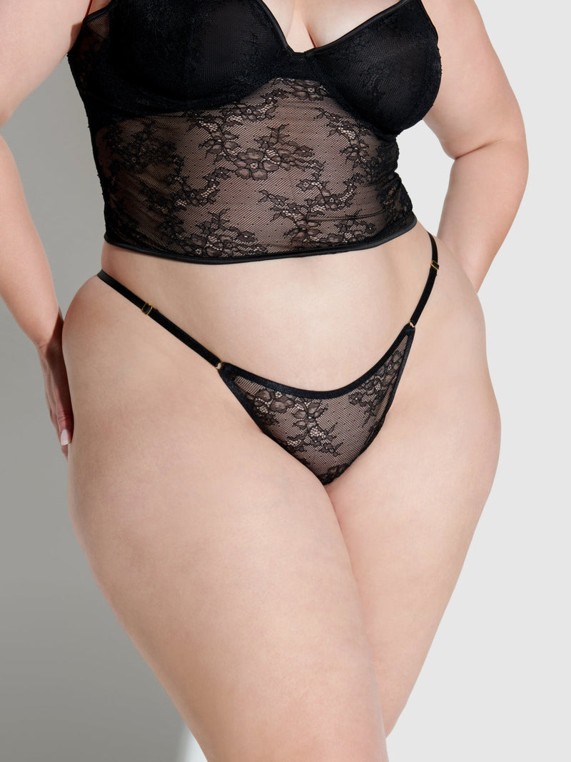 A person models the Frederick's of Hollywood Chiara Lace String Side Bikini, featuring sheer black lace and elastic sides, against a neutral background—celebrating body positivity and plus-size fashion.