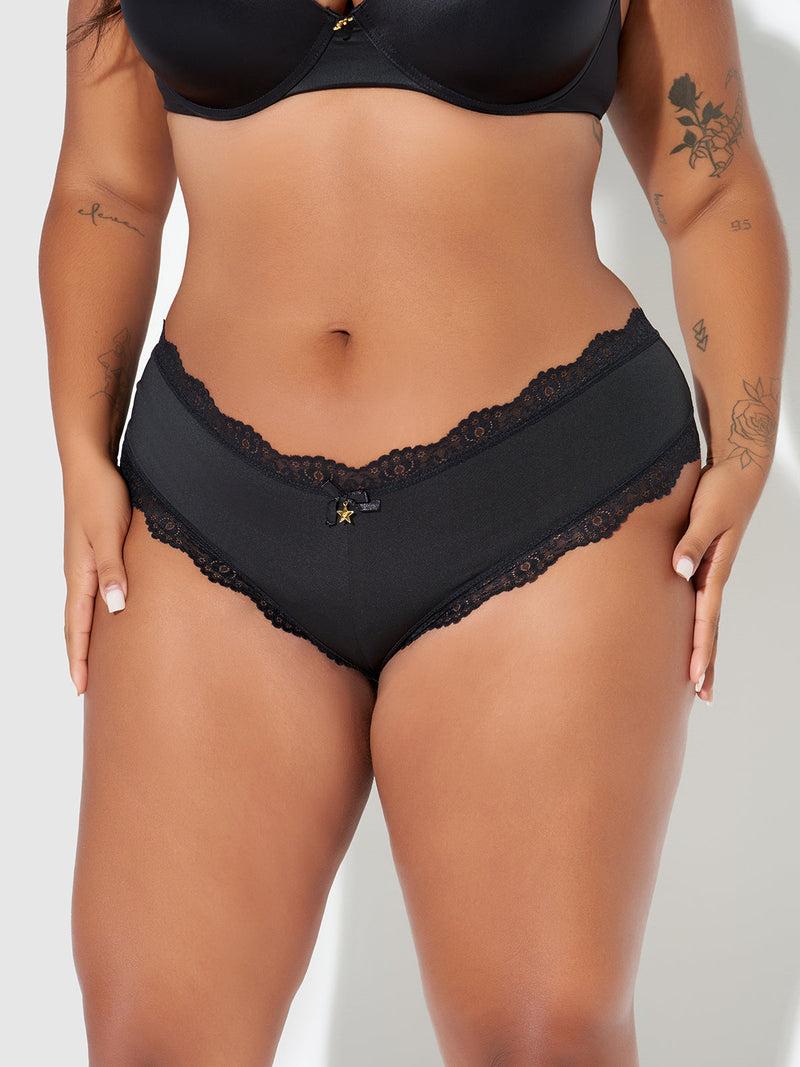 A person poses against a plain backdrop in the Jenna Micro G-String by Frederick's of Hollywood and a black bra, highlighting the silky-soft microfiber lingerie and showing tattoos on their arms and thigh.