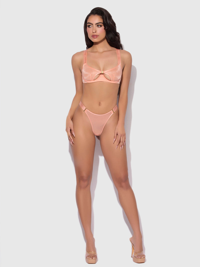 A woman stands against a plain light background wearing the Frederick's of Hollywood Pia Elastic Side Mesh Thong in sheer peach, paired with matching high-waisted bottoms and clear high-heeled shoes. She faces forward, hands at her sides.