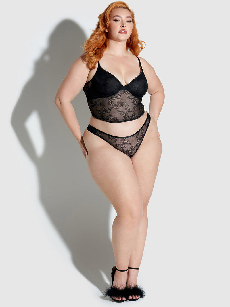 A woman with long red hair models the Chiara Classic Lace Thong by Frederick's of Hollywood, paired with black heels, standing confidently against a plain light gray background.