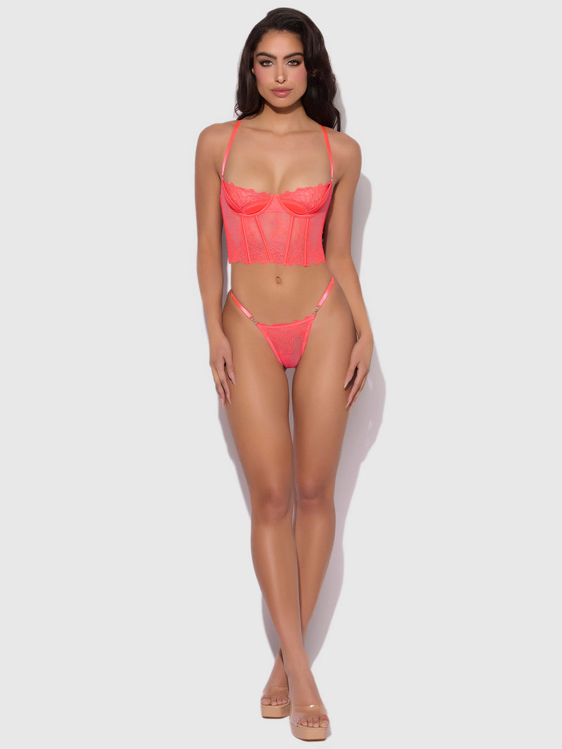 A woman with long dark hair and neutral makeup poses in Frederick's of Hollywood's Wren Embroidered Crotchless Thong With Side Sliders against a white background, styled with a strappy bustier and clear high heels.
