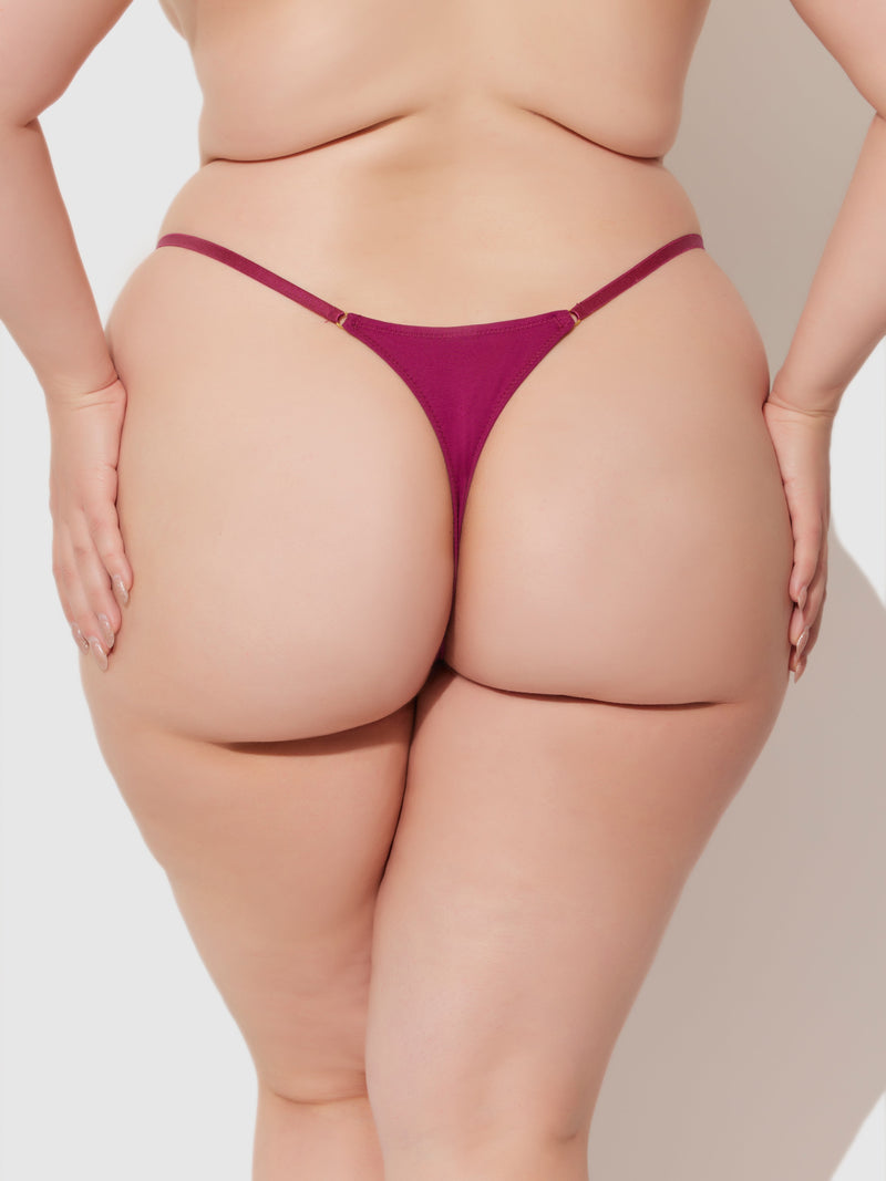 A plus-size person is seen from the back wearing the Bailey Stretch Cotton Thong in burgundy by Frederick's of Hollywood, highlighting natural curves and skin texture with hands on hips against a plain background.