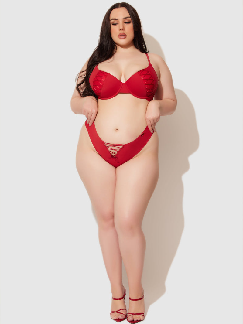 A woman with long dark hair models the Frederick's of Hollywood Natalia Micro Lace Up Front Thong in red, featuring minimal coverage and lace-up details, paired with red heels. She poses against a plain light background, eyes closed and hands pointing downward.