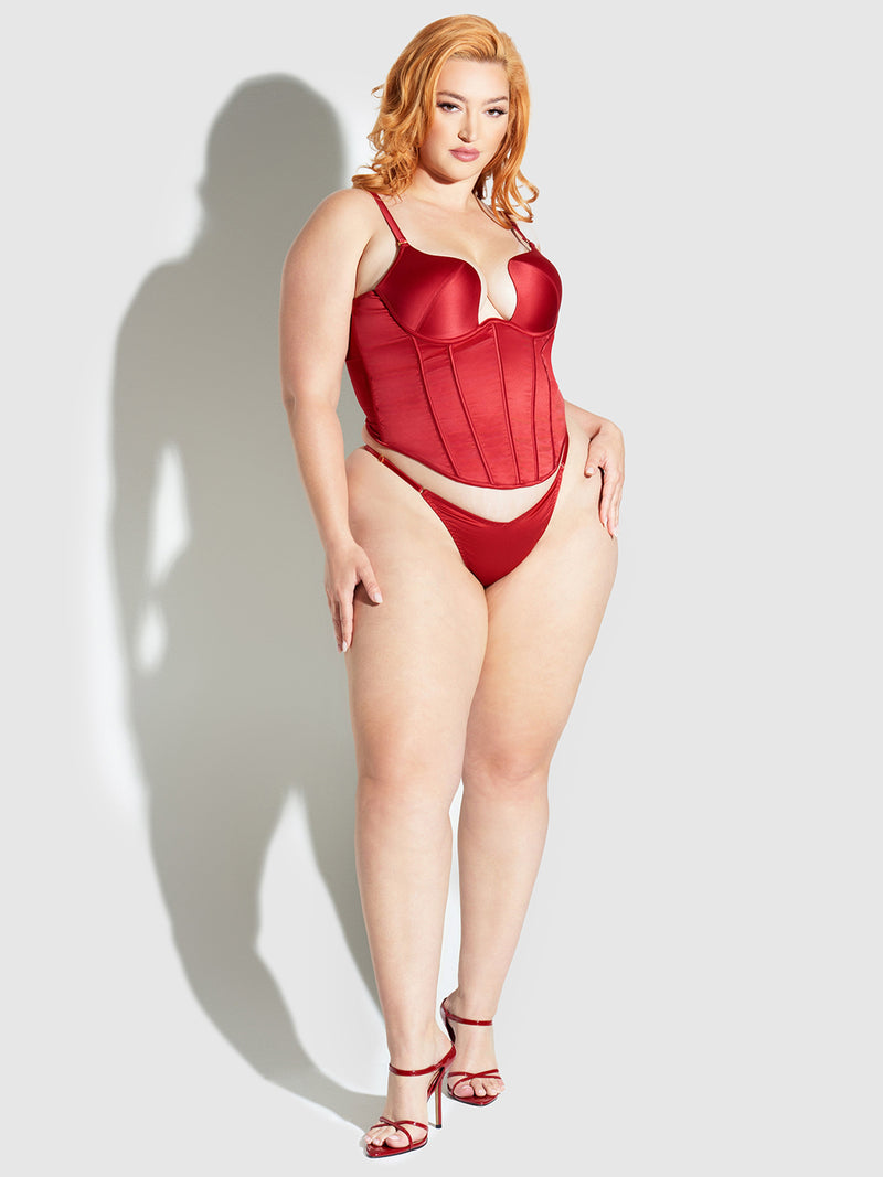 A woman with light skin and red hair poses confidently in a red Frederick's of Hollywood Petra Slider Side Satin Thong, matching satin corset, and red high-heeled sandals against a light gray background.