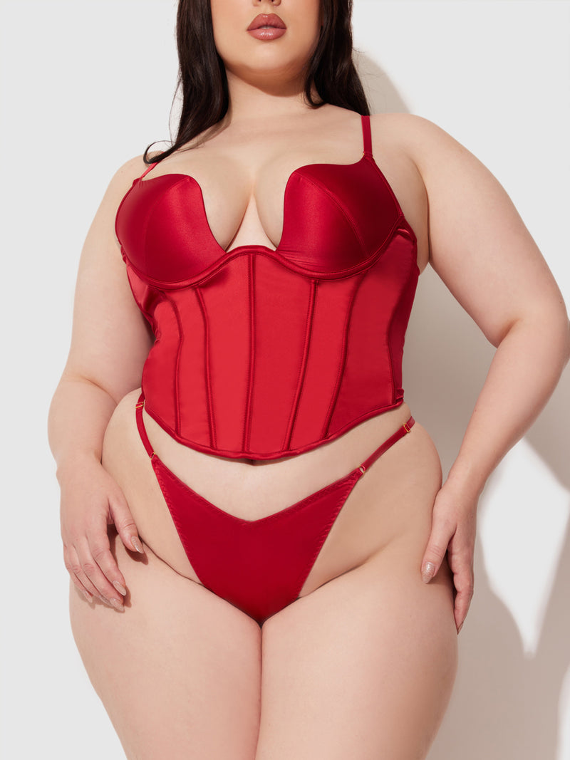 A plus-size woman models the Frederick's of Hollywood Petra Slider Side Satin Thong in red, paired with a matching satin bustier top, posing against a plain white background.