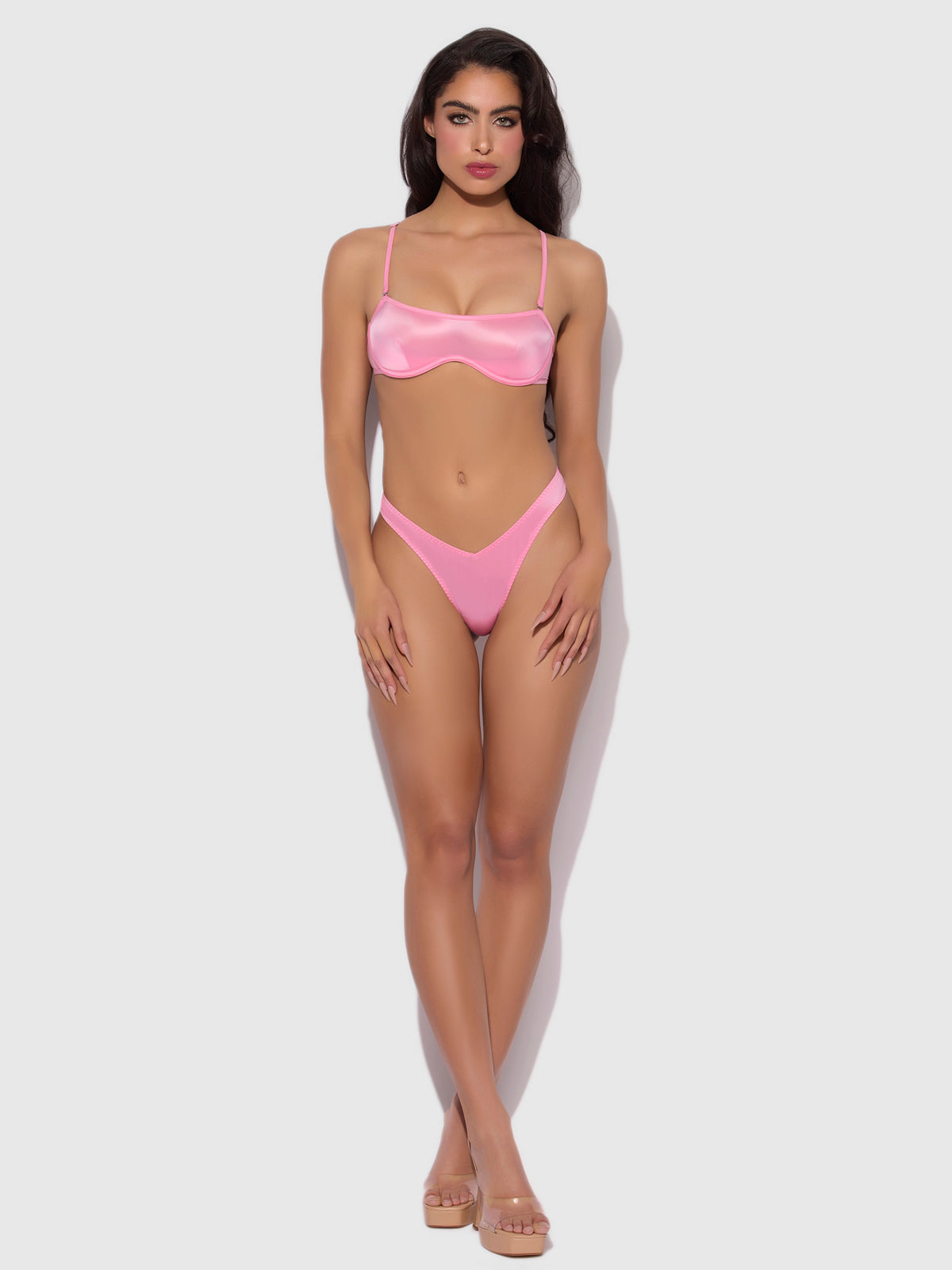 A woman stands on a white background wearing the Frederick's of Hollywood Neev Oil Slick Scoop Thong in shiny pink high-stretch fabric, paired with clear-strap heels, looking directly at the camera.