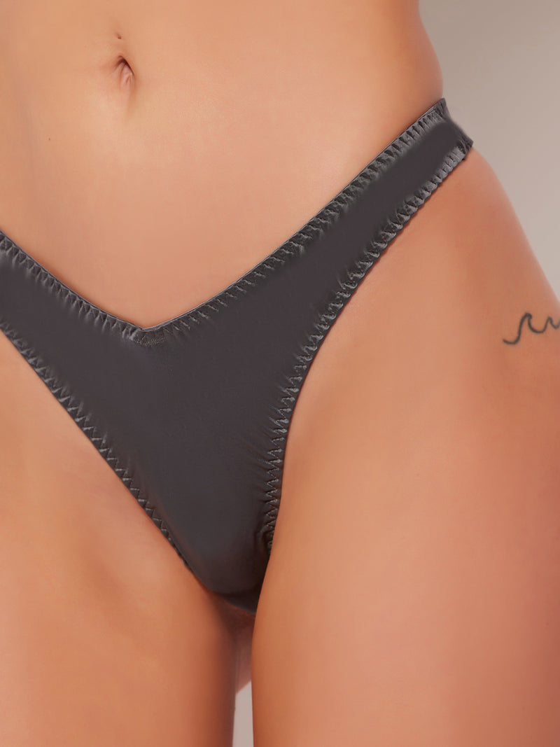 A close-up of a person’s lower torso in Frederick's of Hollywood Neev Oil Slick Scoop Thong, dark gray and high-stretch, with part of their stomach, hip, and a small wave tattoo on the right side visible.