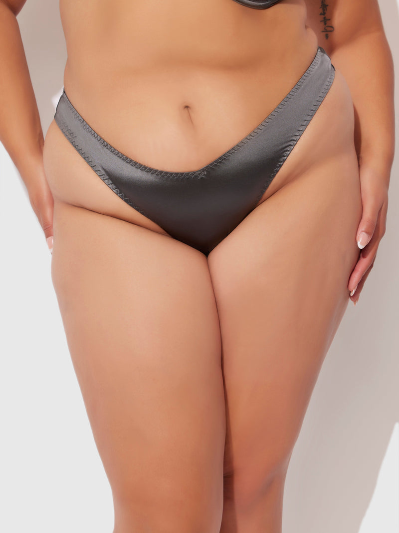 A close-up of a person wearing the Frederick's of Hollywood Neev Oil Slick Scoop Thong in dark gray, standing against a plain light background with manicured hands at their sides.