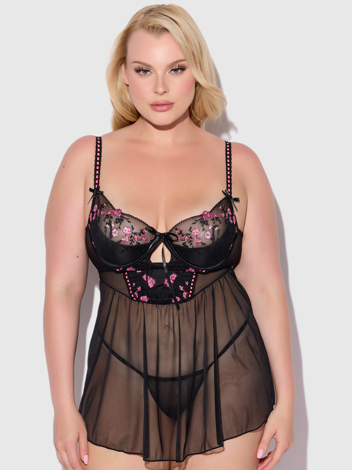 A woman models the Frederick's of Hollywood Harlow Embroidered Mesh & Elastic Longline Babydoll, featuring sheer black fabric with pink floral embroidery, matching underwear, and strappy black bows against a plain light background.