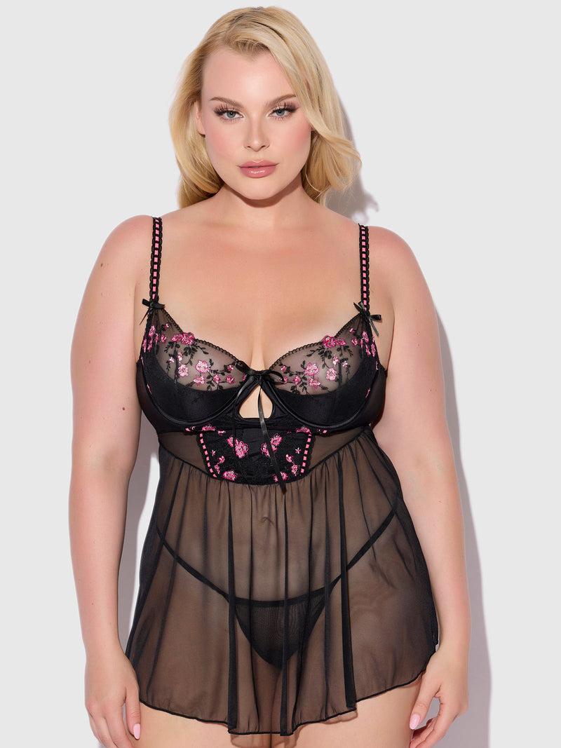 A woman models the Frederick's of Hollywood Harlow Embroidered Mesh & Elastic Longline Babydoll, featuring sheer black fabric with pink floral embroidery, matching underwear, and strappy black bows against a plain light background.