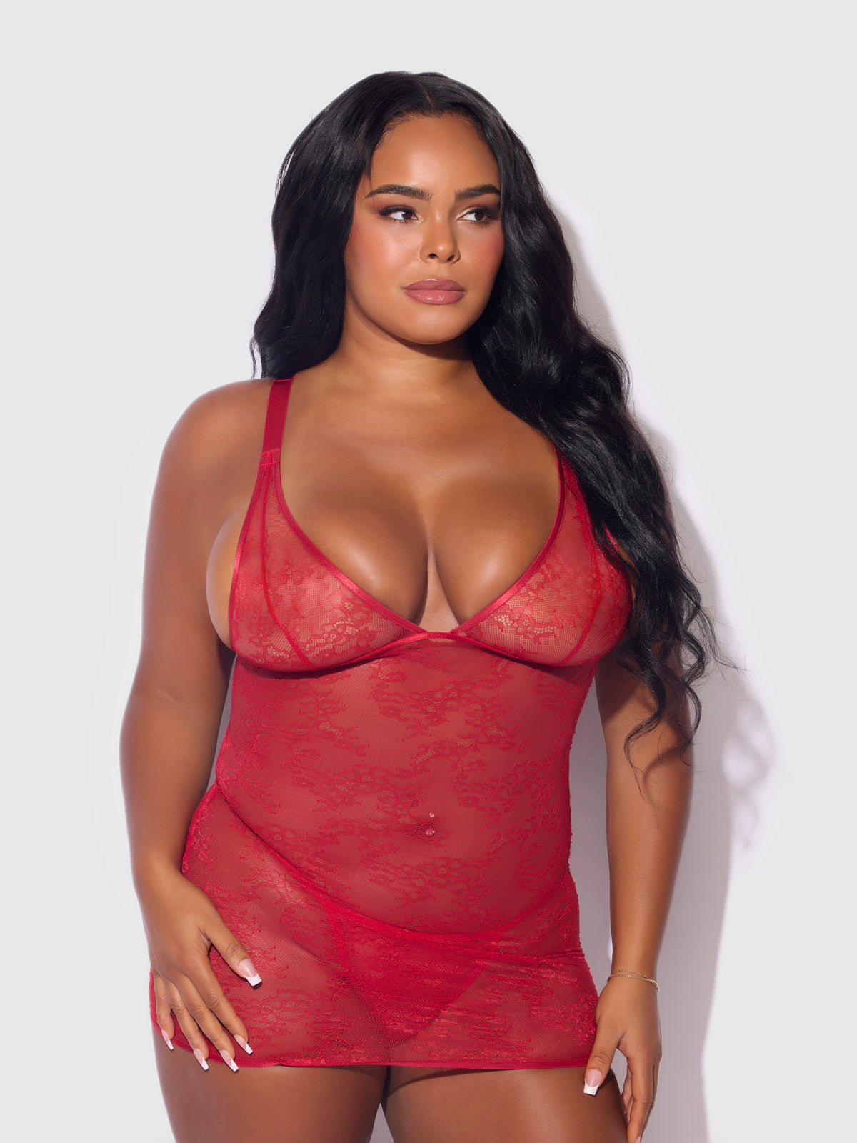 A woman with long, wavy black hair wears the Frederick's of Hollywood Chiara Lace & Strappy Open Back Chemise, featuring sheer red lace and thin straps, standing against a plain light background.