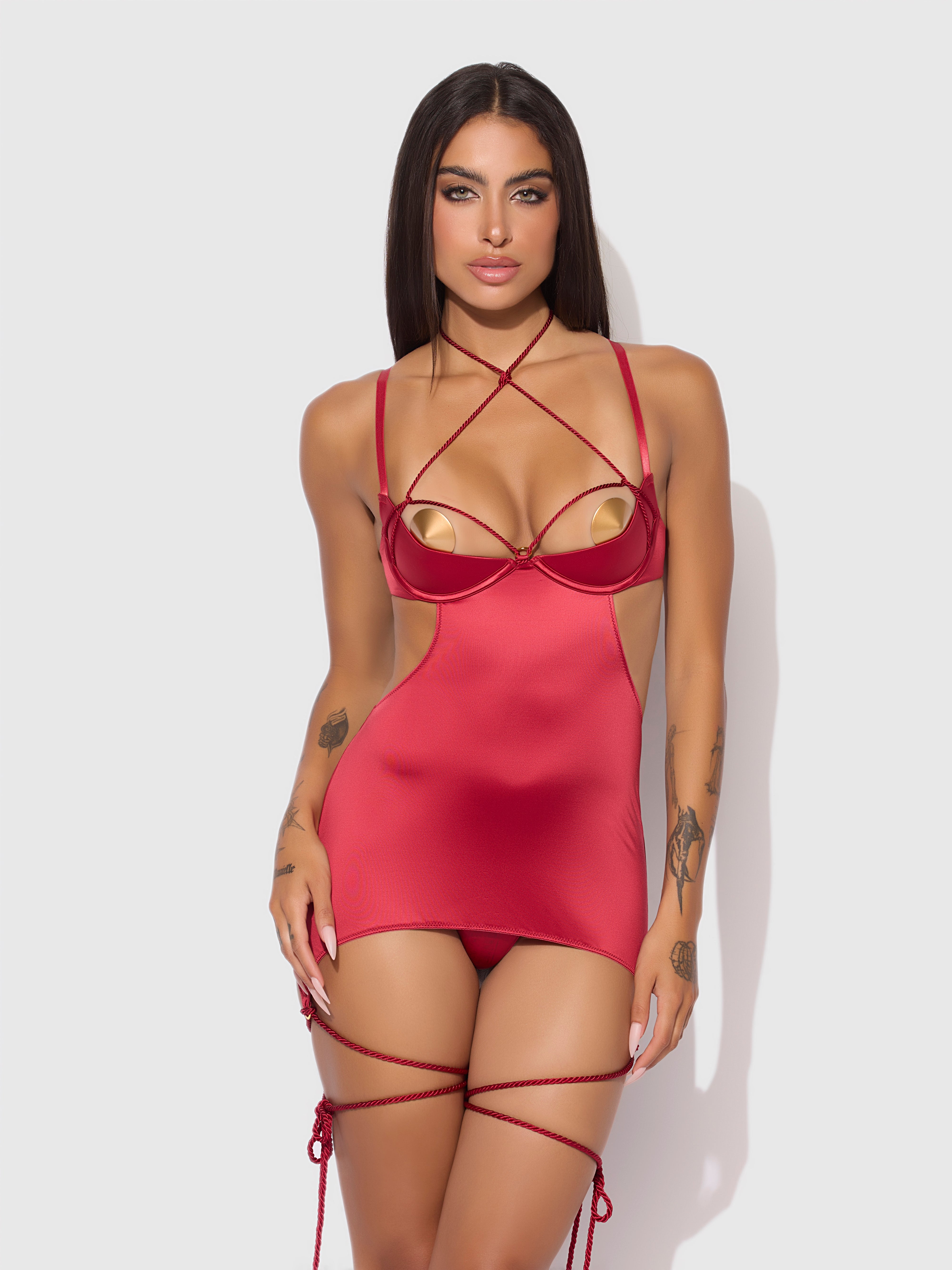 A woman with long dark hair models the Frederick's of Hollywood Nadya Liquid Satin & Rope Open Back Chemise & G-String Set, featuring gold cups and matching rope thigh garters, against a plain light background.