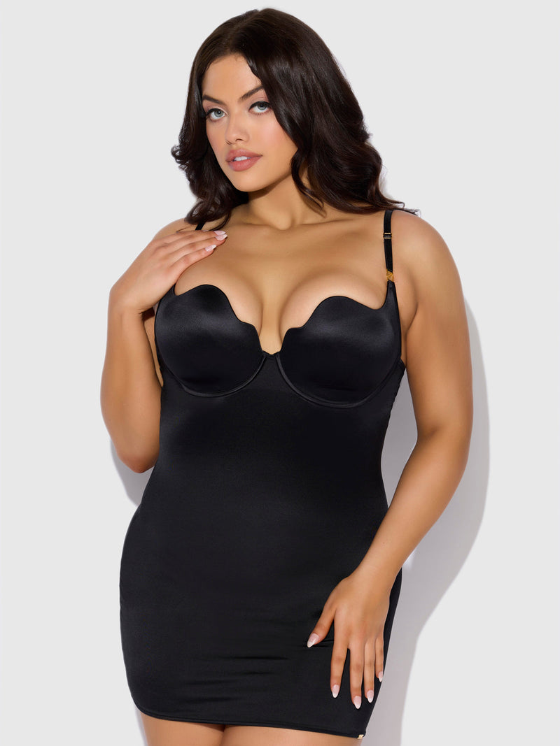 A woman wears the Director's Cut Liquid Satin Chemise by Frederick's of Hollywood—a fitted black mini dress with a sweetheart neckline—standing against a light background, touching her shoulder with one hand.