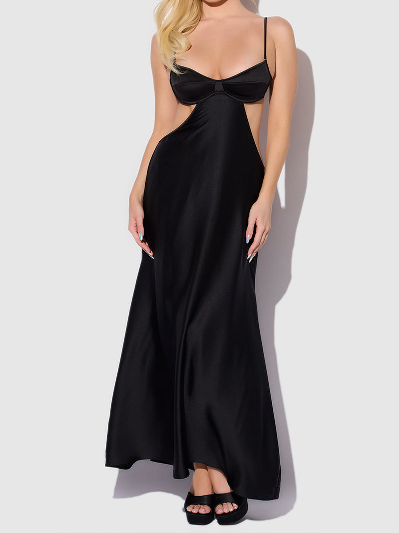 A woman with blonde hair stands against a white background, wearing the Petra Satin Monowire Cutout Gown by Frederick's of Hollywood—a long black gown featuring thin straps, a supported fit with monowire cutouts, and an open back. She completes the look with black open-toe heeled sandals.
