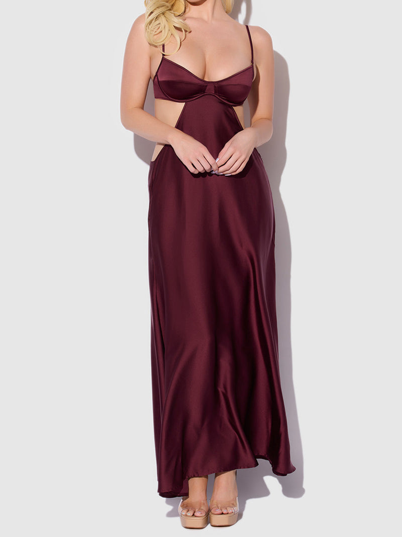A woman stands against a plain light background wearing the Frederick's of Hollywood Petra Satin Monowire Cutout Gown—a long, burgundy satin gown featuring spaghetti straps, a supported monowire fit, cut-out sides, and a deep neckline.