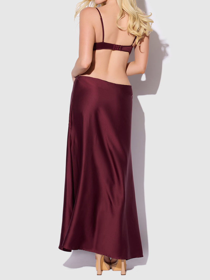 A woman with long blonde hair is shown from the back, wearing the Frederick's of Hollywood Petra Satin Monowire Cutout Gown in dark burgundy. The gown features a satin monowire for a supported fit and cutout detailing. She stands against a plain light background, paired with nude heels.