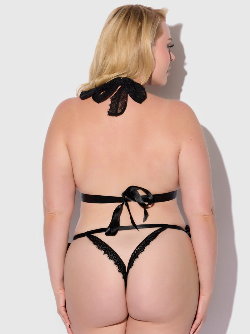 A woman with light skin and blonde hair stands with her back turned in the Frederick's of Hollywood Rosalind Satin & Lace Drop Off Crotchless Teddy, featuring black lace, ribbon neck and back ties, an open back, and set against a plain light background.