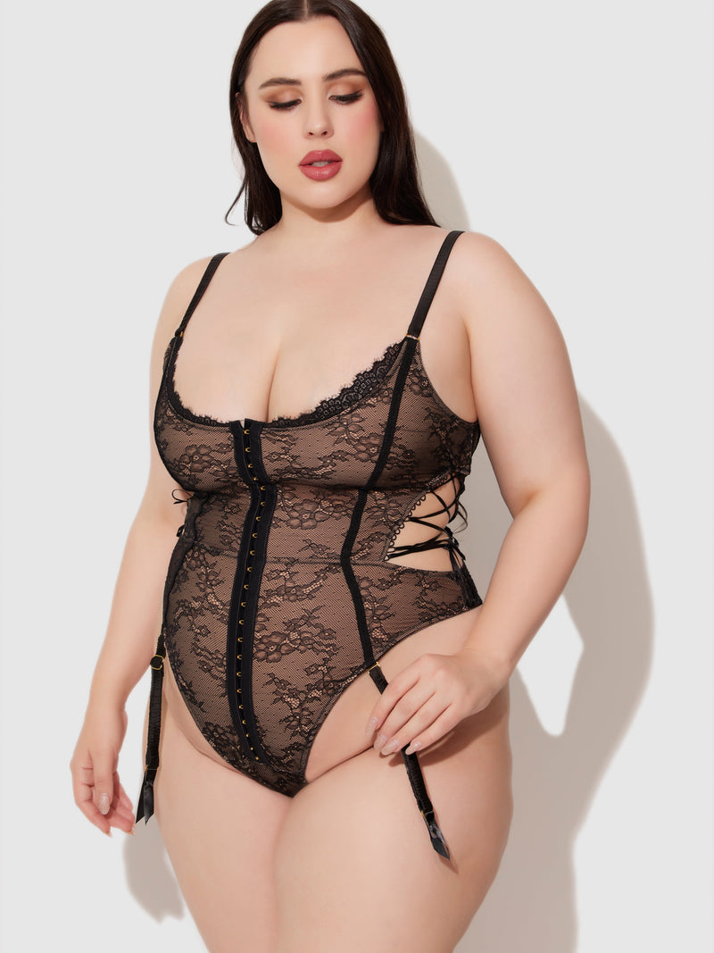 A woman in the Luella Hook & Eye Front Strappy Lace Teddy by Frederick's of Hollywood, featuring lace-up sides and adjustable straps, stands against a plain light background, looking down with a relaxed expression.