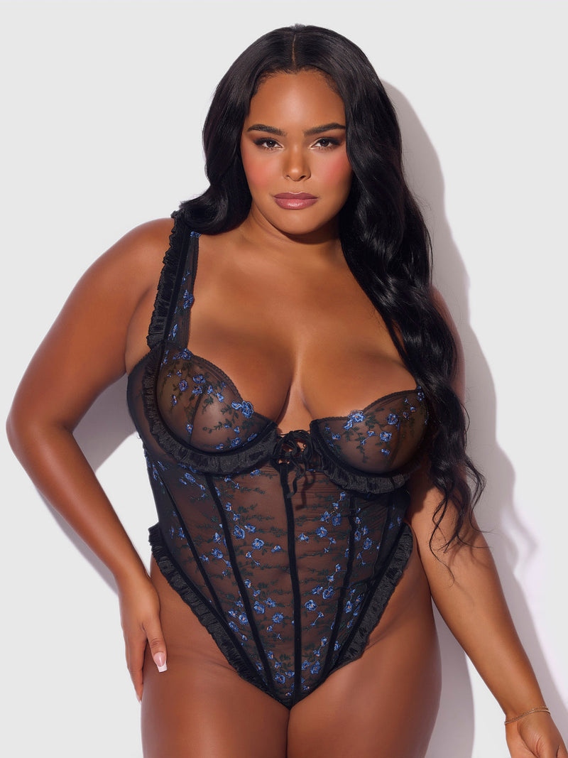 A woman with long, wavy dark hair models the Frederick's of Hollywood Harlow Embroidered Stretch Mesh Teddy, a sheer black lace bodysuit with blue floral embroidery, posing confidently against a light gray background.