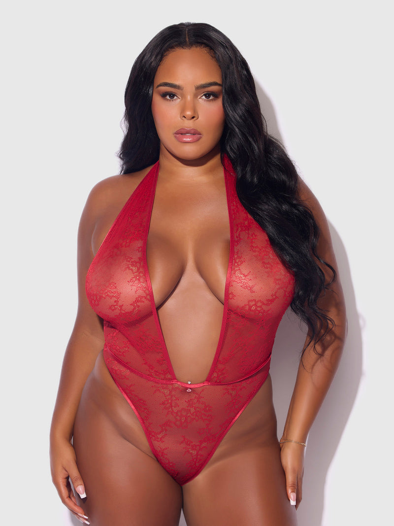 A woman with long, wavy black hair models the Chiara Crotchless Halter Lace Teddy by Frederick's of Hollywood, featuring sheer floral lace and a deep V-neck, standing against a plain light background.