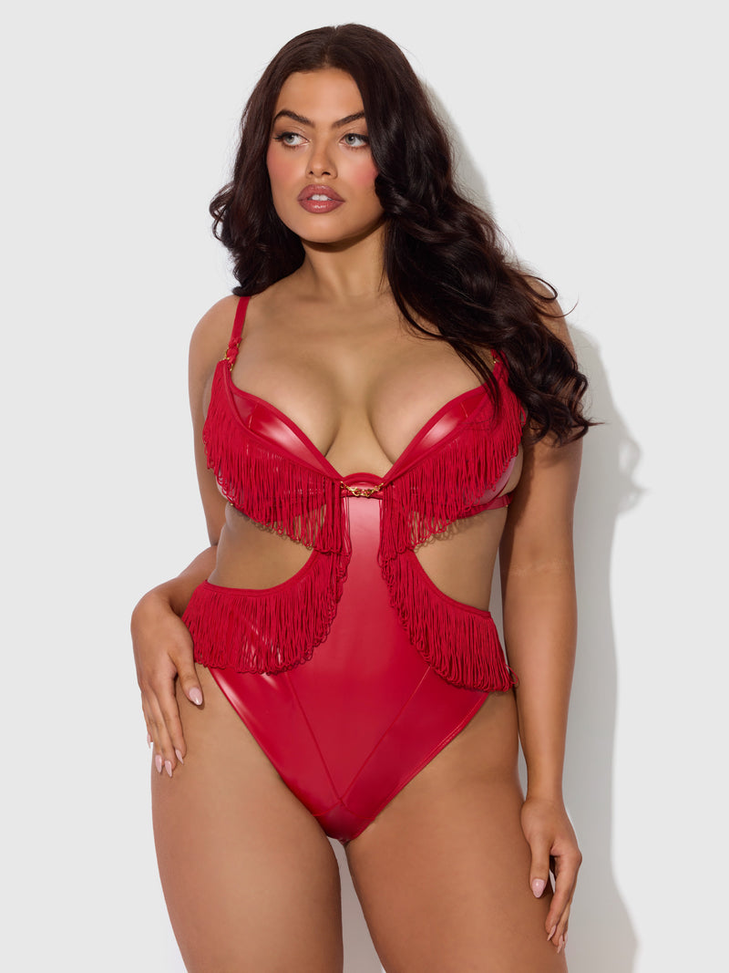 A woman with long dark hair models the Frederick's of Hollywood Hendrix Faux Leather & Fringe Crotchless Teddy, featuring red faux leather, fringe details, and cut-out sides, standing against a light gray background.