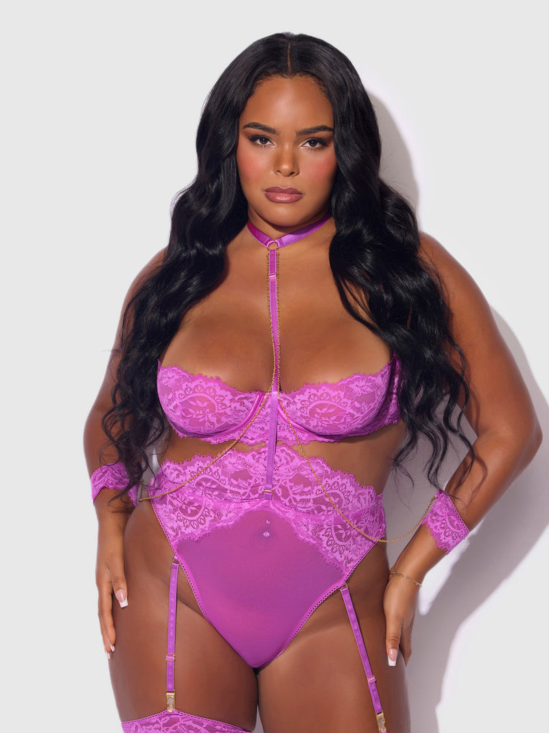 A woman with long wavy black hair confidently poses in the Frederick's of Hollywood Arianna Lace & Chains Thigh Garter Teddy, featuring harness accents and matching wrist accessories, against a plain light background.