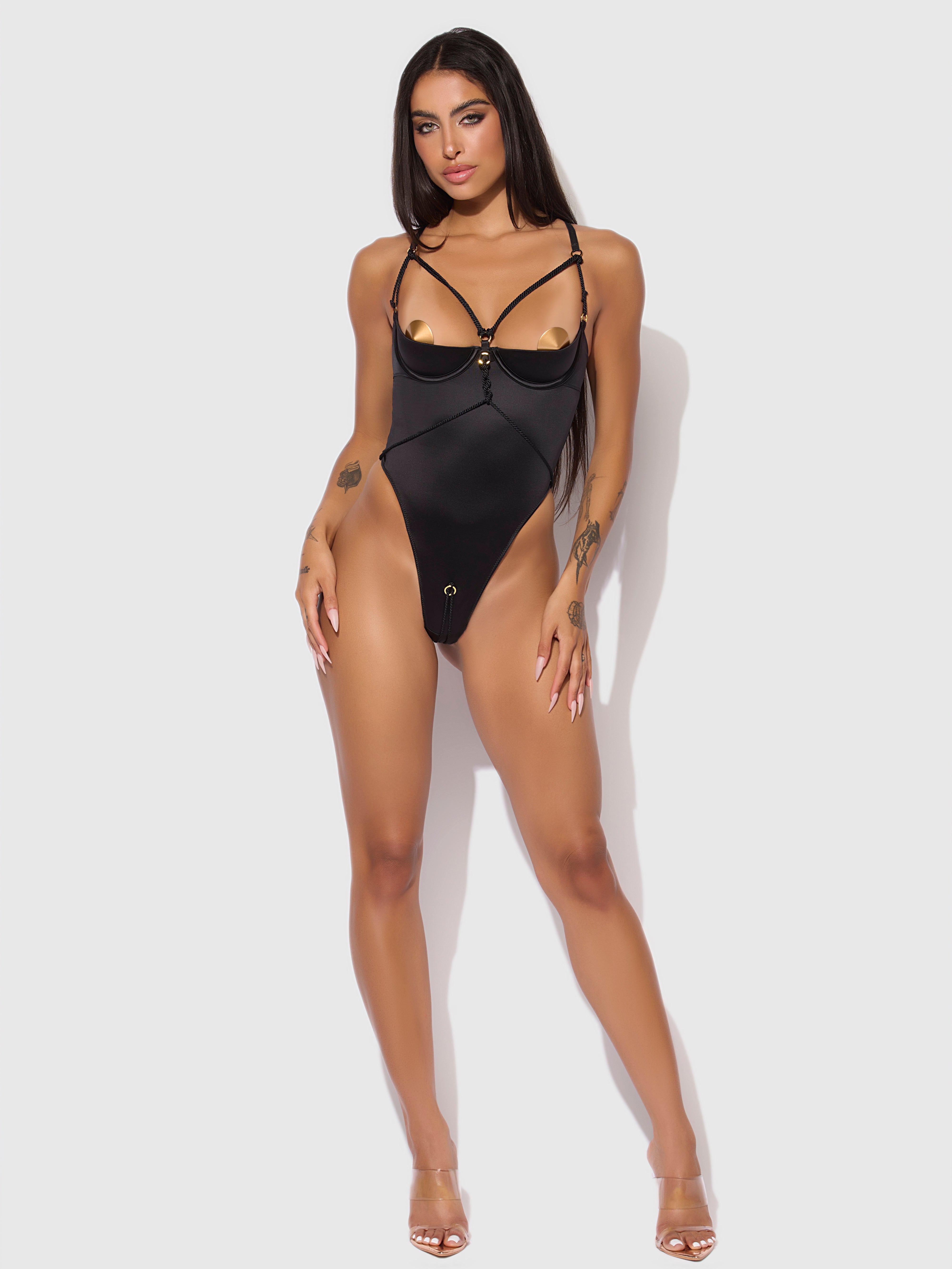 A woman with long dark hair and visible tattoos poses confidently against a white background wearing the Frederick's of Hollywood Nadya Liquid Satin & Rope Crotchless Teddy and clear high heels.