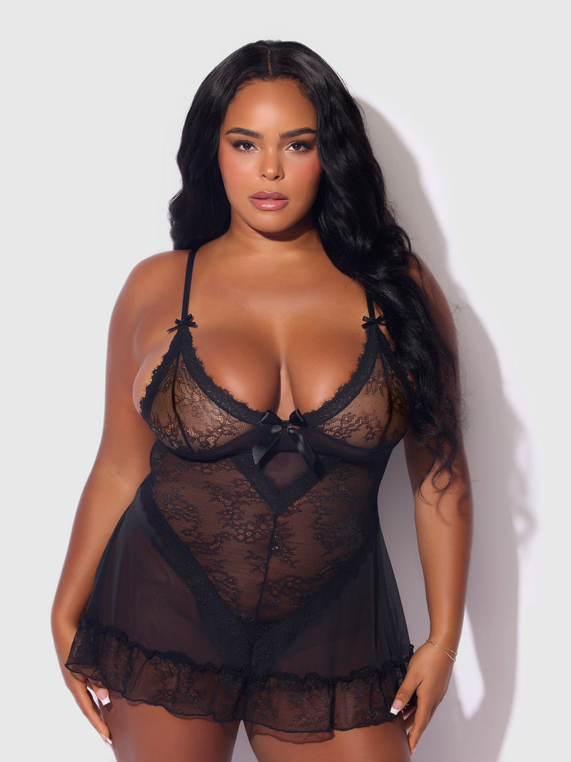 A woman with long, wavy black hair poses confidently in the Frederick's of Hollywood Chiara Lace & Mesh Babydoll Set, featuring sheer mesh, ribbon details, and delicate stretch lace against a plain light background.