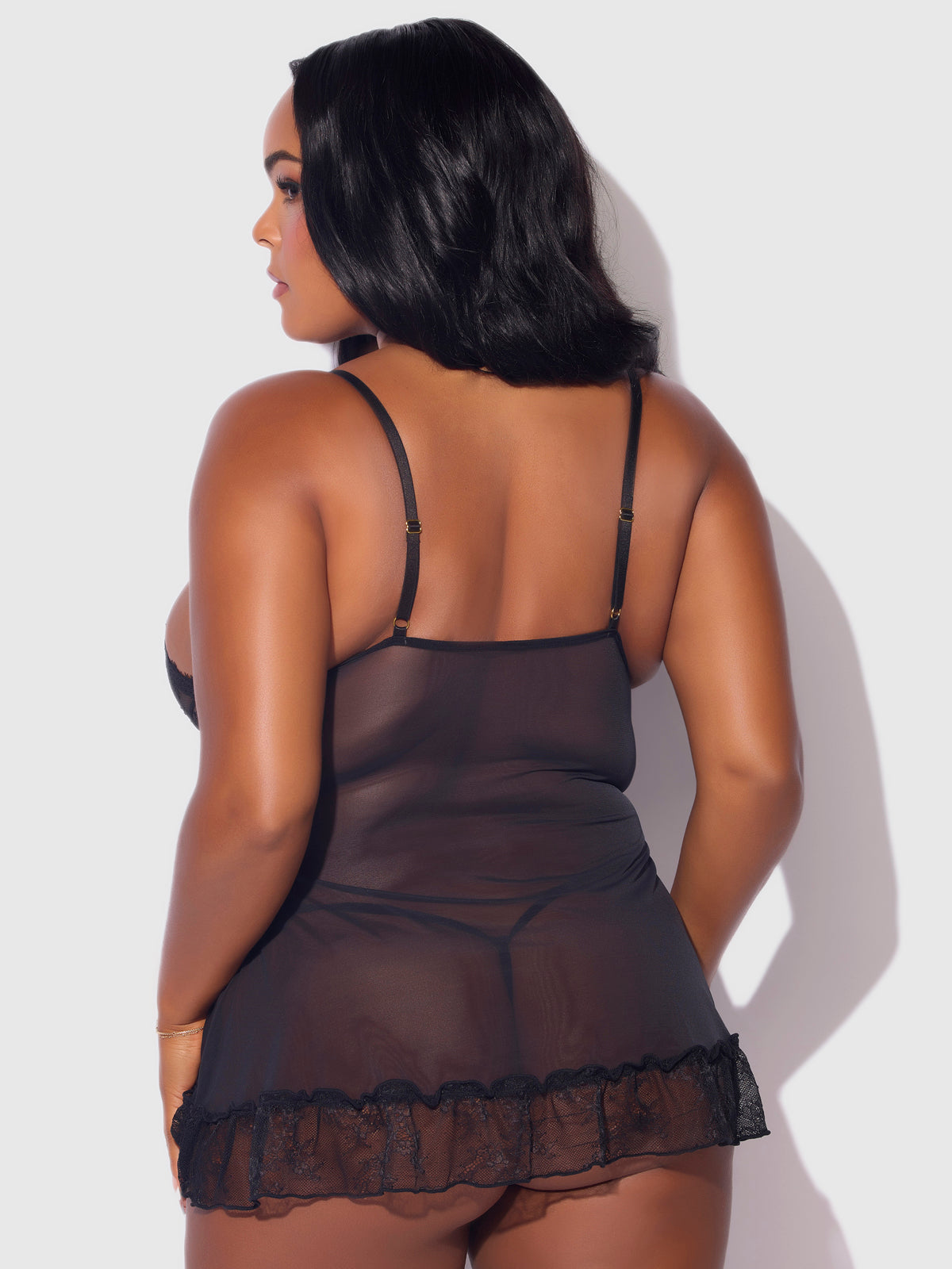 A woman with long dark hair is shown from the back, wearing the Frederick's of Hollywood Chiara Lace & Mesh Babydoll Set, featuring sheer black mesh, adjustable straps, and a ruffled lace hem, against a plain light background.