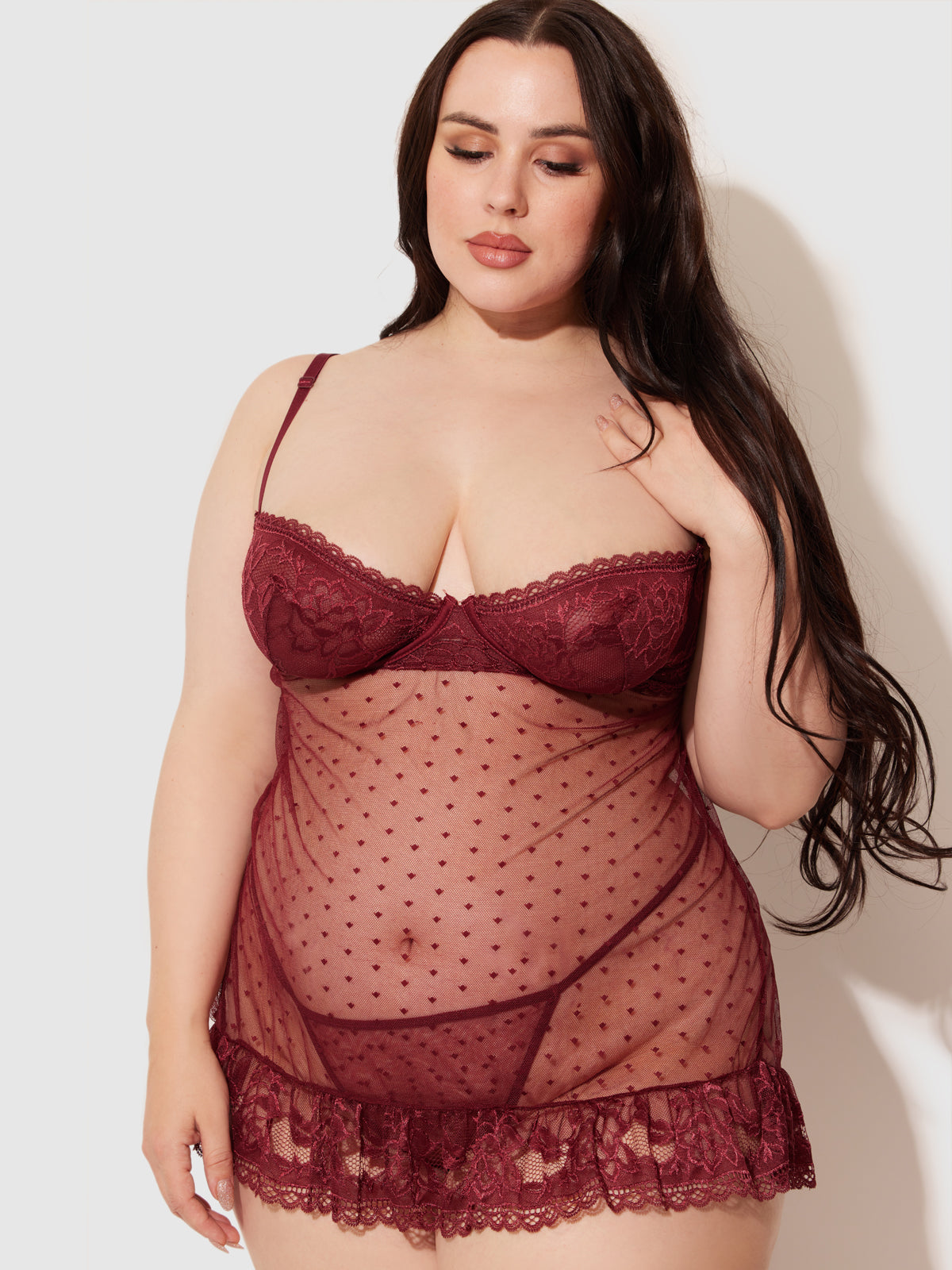 A woman with long dark hair models the Riley Lace Babydoll Set by Frederick's of Hollywood, featuring burgundy lace, sheer swiss dot mesh, and polka dot details against a light background.